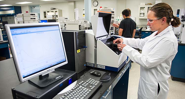 26487 Photo of a woman in a white lab coat and safety glasses placing samples for testing into a small, box-like piece of equipment; and next to the equipment is a computer.