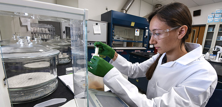 feedstocks-51829 A woman in safety glasses, rubber gloves, and lab coat measures a sample in a laboratory.