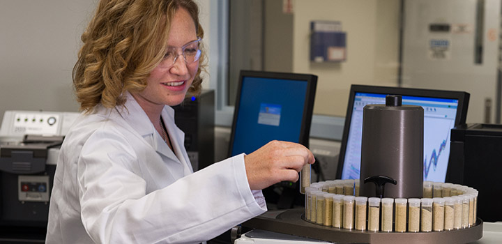 nir-34510 A woman in a lab coat and safety glasses looks at a vial in a laboratory.