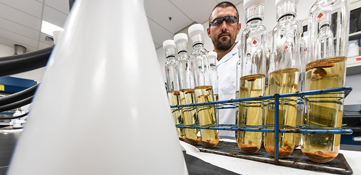 slurries-51835 A man in safety glasses and lab coat stands behind a row of glass vials with yellow liquid in a laboratory.
