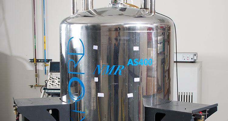 tcuf-19989 Photo of a 400 MHz Agilent Unity Inova NMR Spectrometer, which is composed of a large metal tank on a three-legged metal stand; clear plastic tubes and wires stick out of the top and bottom of the tank.