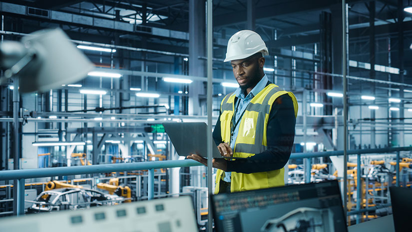neb-manufacturing-worker Worker stands inside a manufacturing plant overlooking laptop