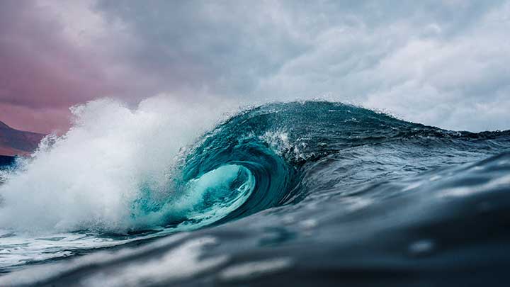 Photo of a crashing ocean wave, pink and blue clouds in the background. 
