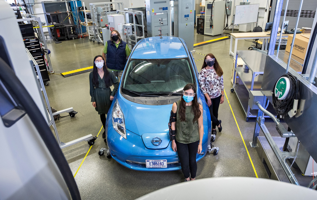 Four people wearing masks stand in a circle around an electric vehicle.